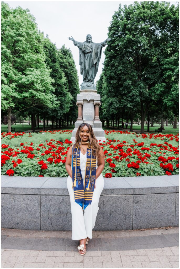 Graduate posing in front of a statue and red flowers photographed by Leah Rife Photo on the University of Notre Dame campus.