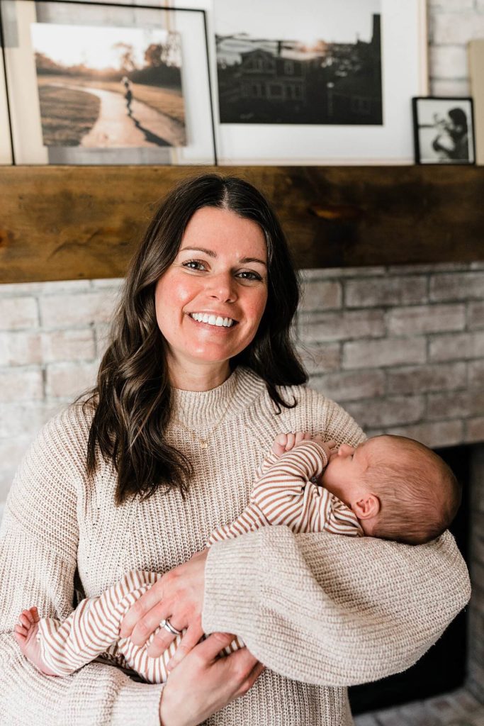 mother holds newborn in front of fireplace during in-home newborn photography session in Carmel Indiana